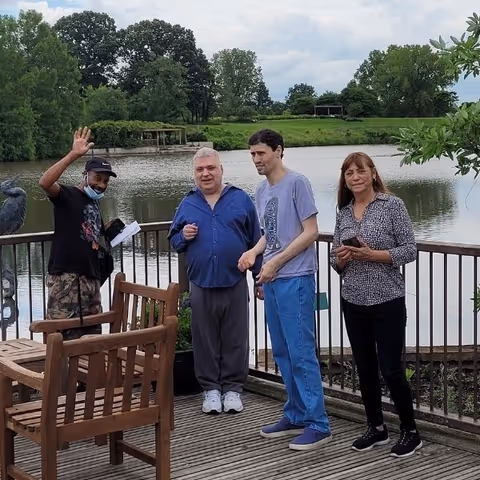 Four people stand on a wooden deck by a lake, near lush trees.