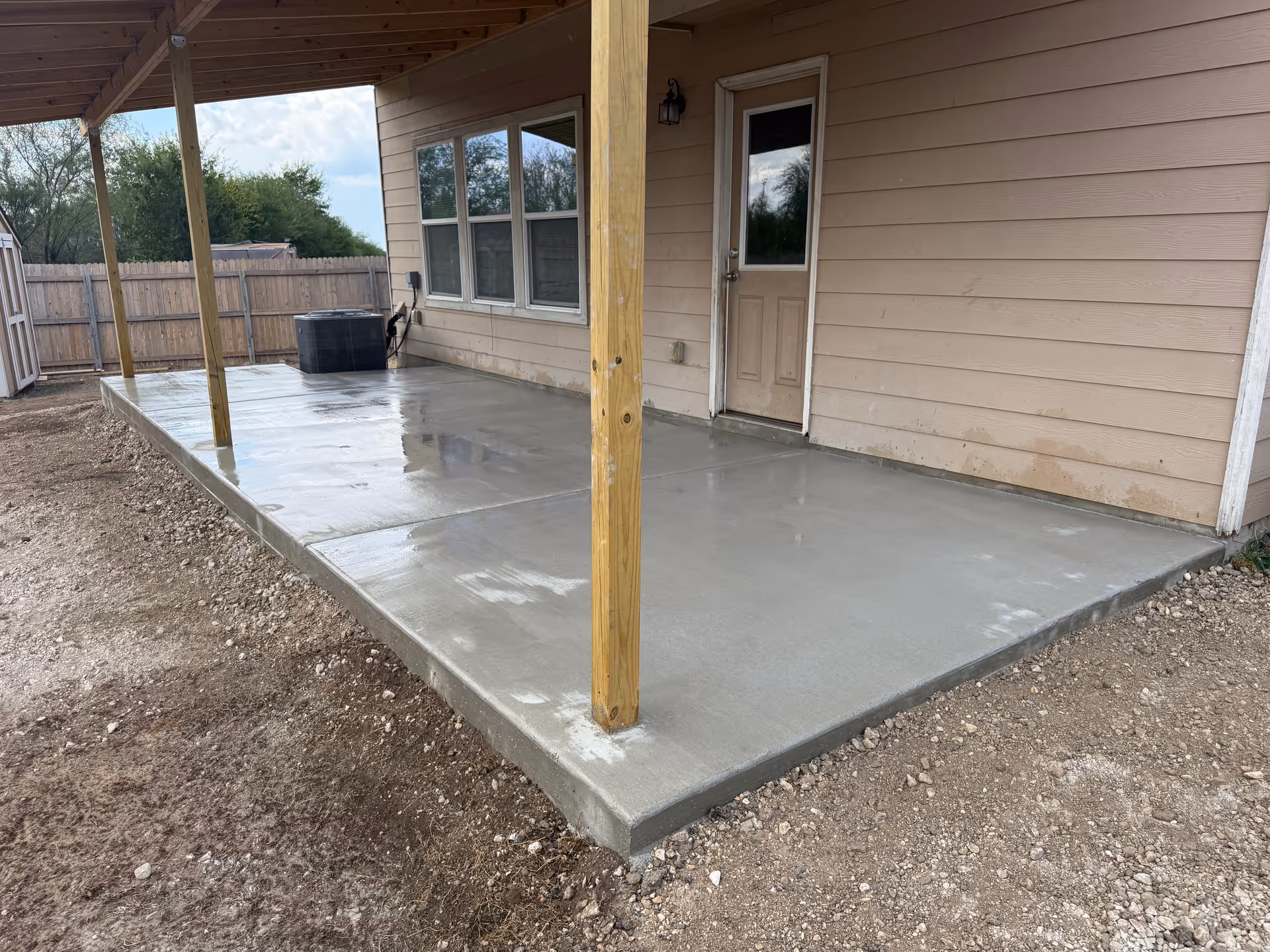 A covered concrete patio attached to a beige house with a door and windows.