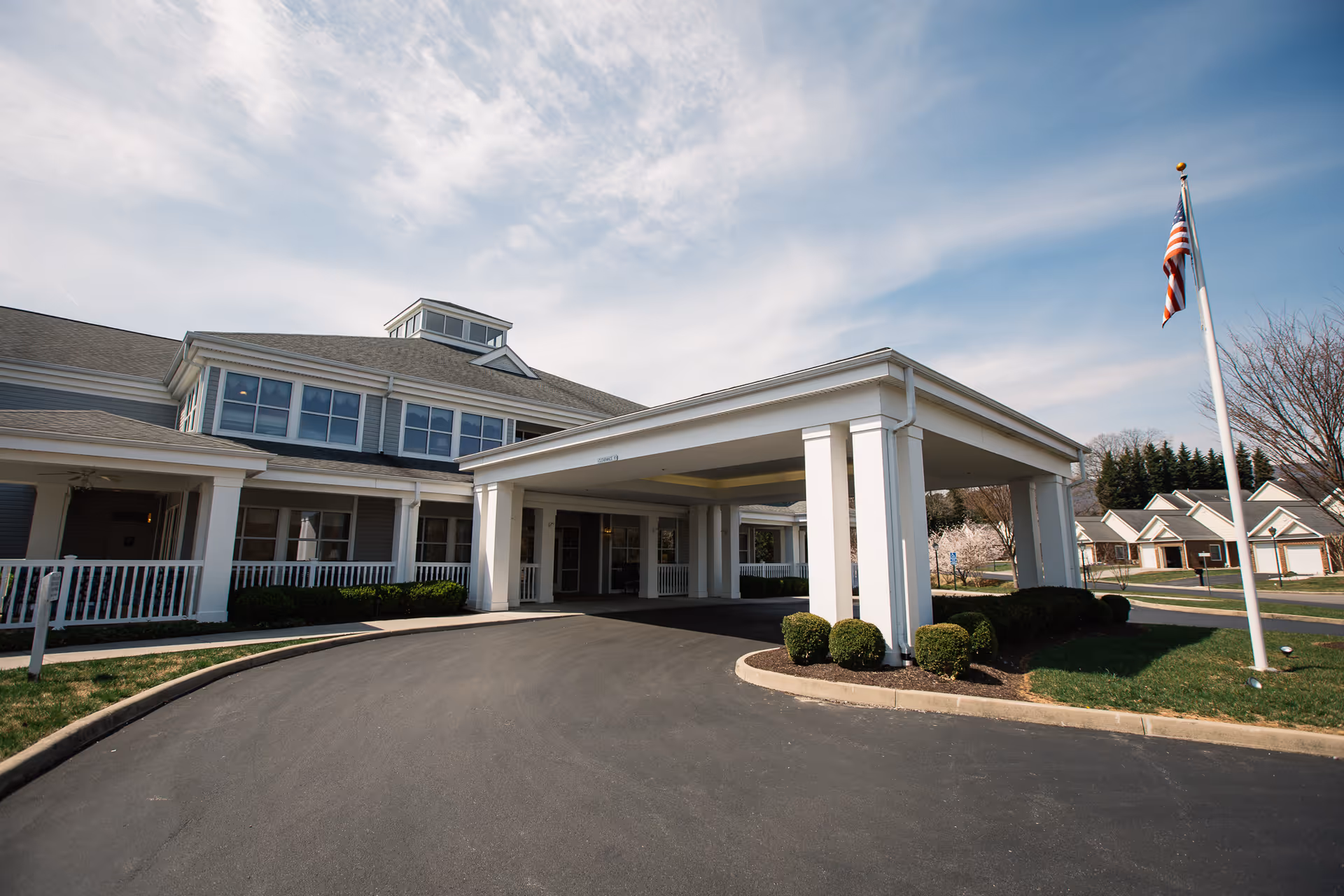 Exterior view of a senior living facility named Friendship Salem Terrace, showing the main entrance with a covered driveway, white columns, and an American flag on a flagpole. The building has multiple windows and a well-maintained lawn with bushes and trees under a partly cloudy sky.