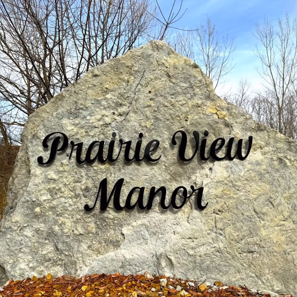 Large rock sign reads Prairie View Manor with leaf-covered ground.