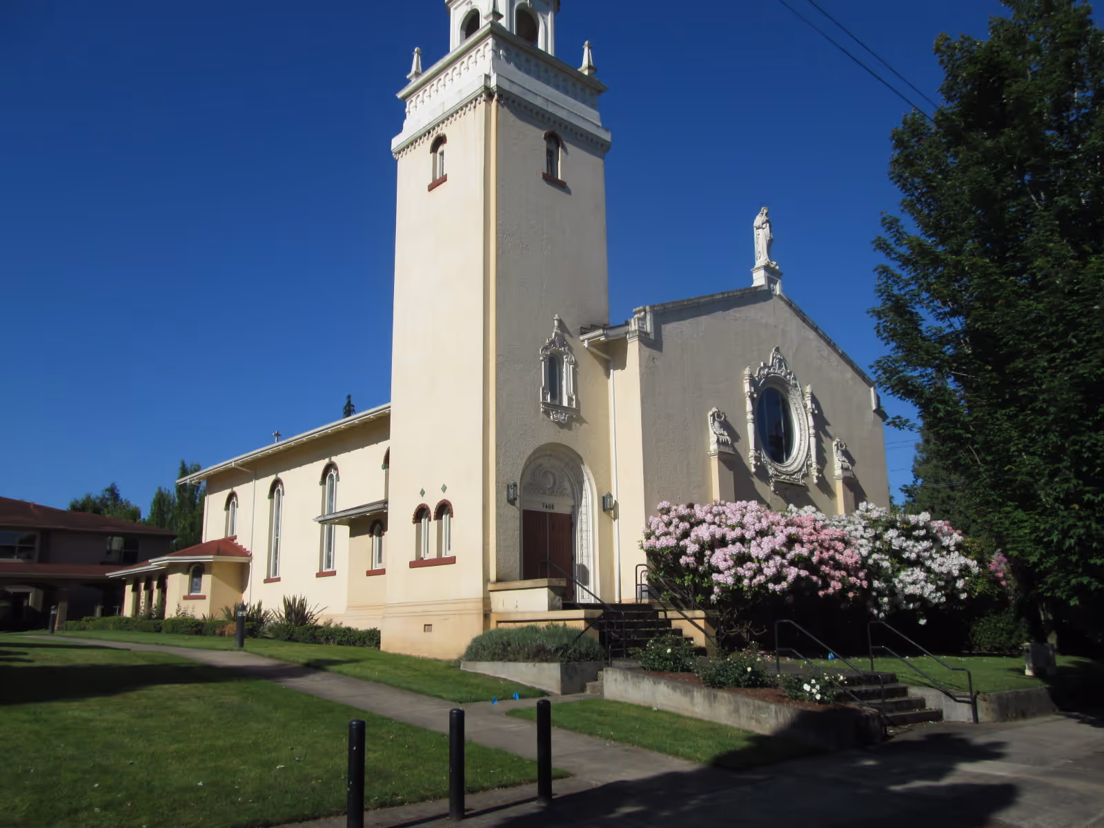 Assumption Village building with a tall tower and blooming shrubs.
