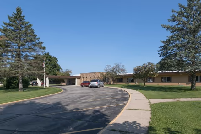 Exterior view of a single-story building with a curved driveway and two parked cars in front. The building is surrounded by green grass, trees, and a clear blue sky.