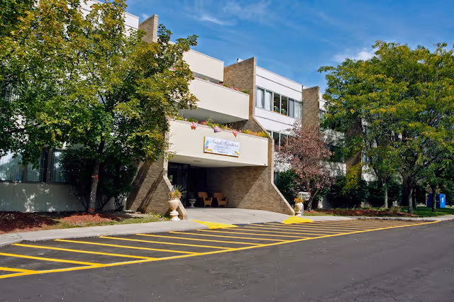 Exterior view of Angel Gardens Assisted Living facility showing the entrance with a covered drop-off area, surrounded by trees and landscaping under a blue sky.