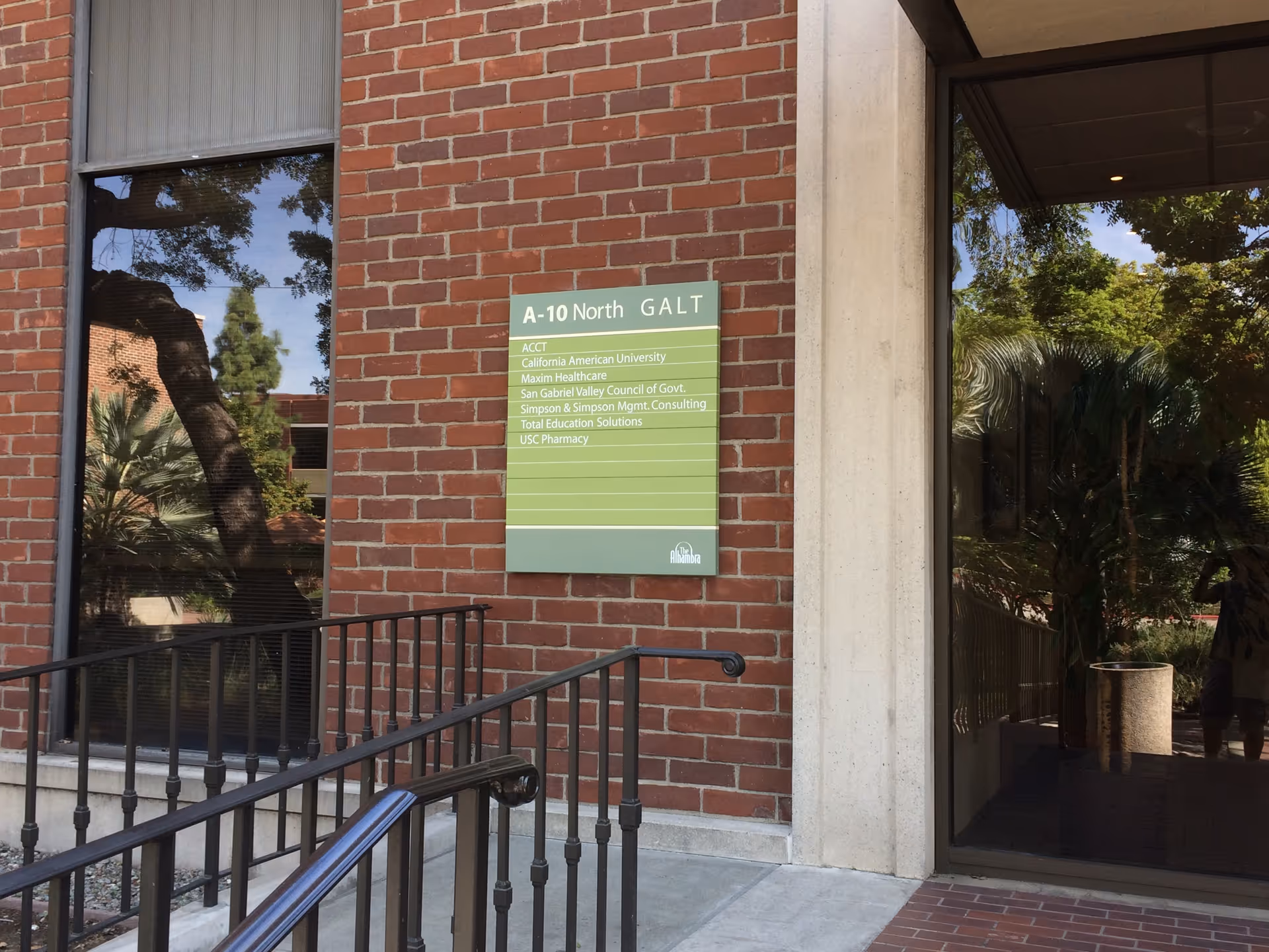Brick building entrance with green directory sign beside glass doors and railing.