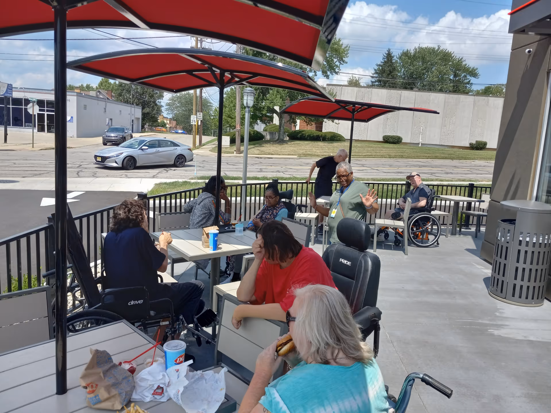 Several elderly individuals, some in wheelchairs, sitting and socializing at outdoor tables under red umbrellas on a patio area next to a street with cars and buildings in the background.