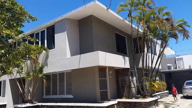 Exterior view of a two-story building with gray walls and multiple windows, surrounded by palm trees and other greenery under a clear blue sky. A person in a red shirt is walking near a parked silver car on the right side.