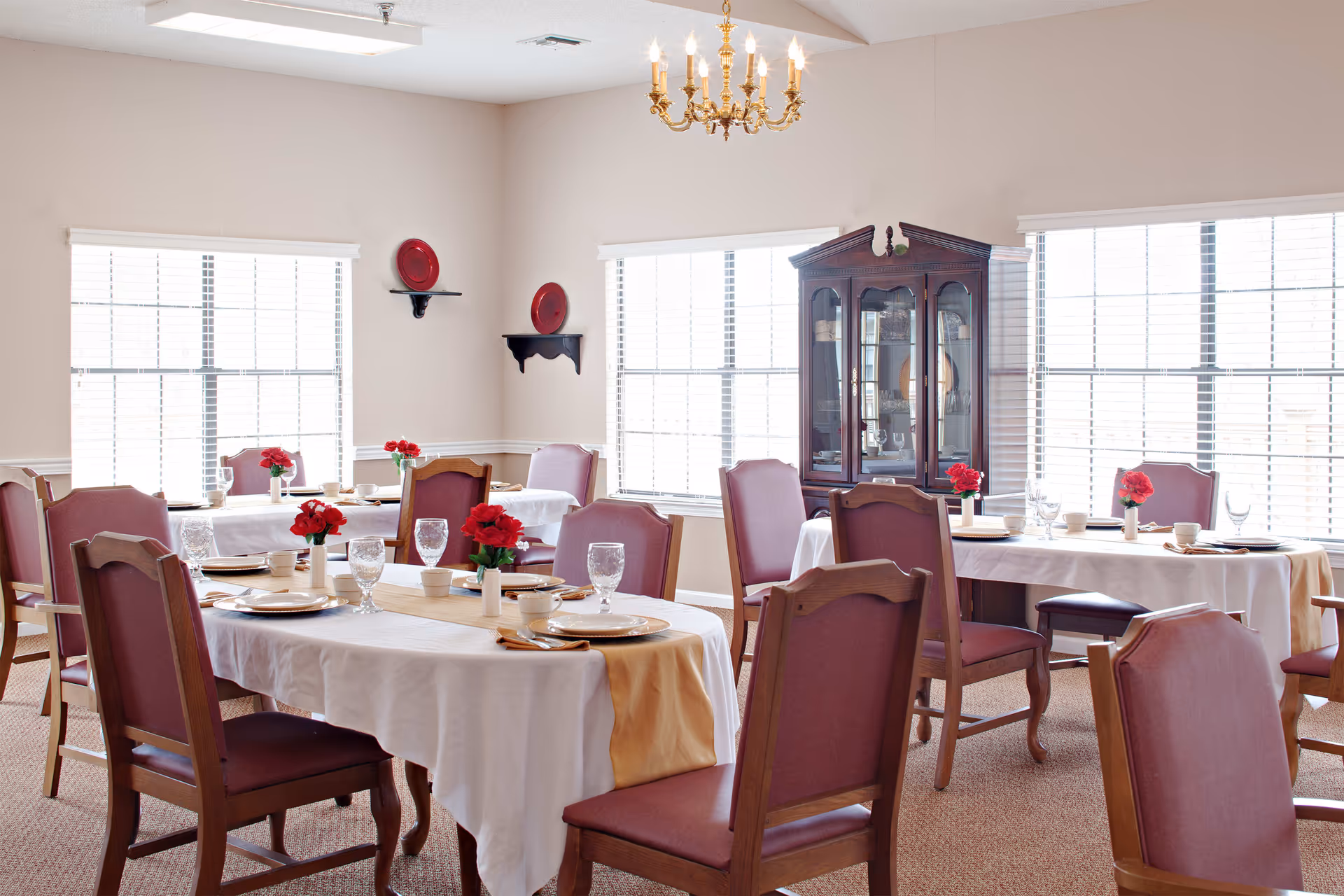 A bright dining room with several tables covered in white tablecloths and gold runners, each set with plates, glasses, and red flower centerpieces. The room has large windows with white blinds, a wooden china cabinet, and a chandelier hanging from the ceiling.