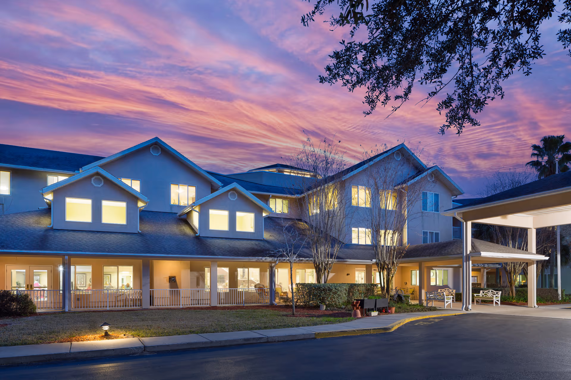 Exterior view of Augustine Landing senior living facility at dusk with illuminated windows, a covered entrance, benches, and a vibrant purple and pink sky in the background.