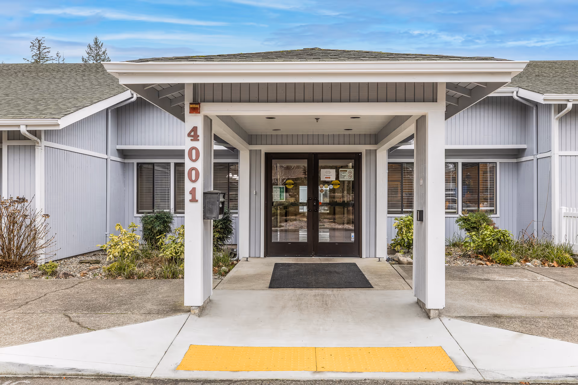 Entrance of a building with the number 4001 displayed on a white pillar. The building has light gray siding, a covered entryway with double glass doors, and some landscaping with bushes on either side of the entrance.