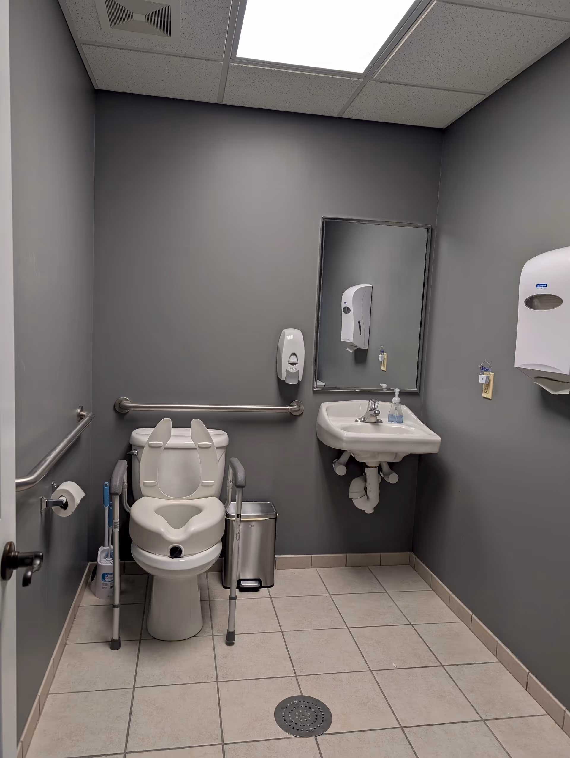 Accessible bathroom with toilet, sink, grab bars, and wall-mounted dispensers.