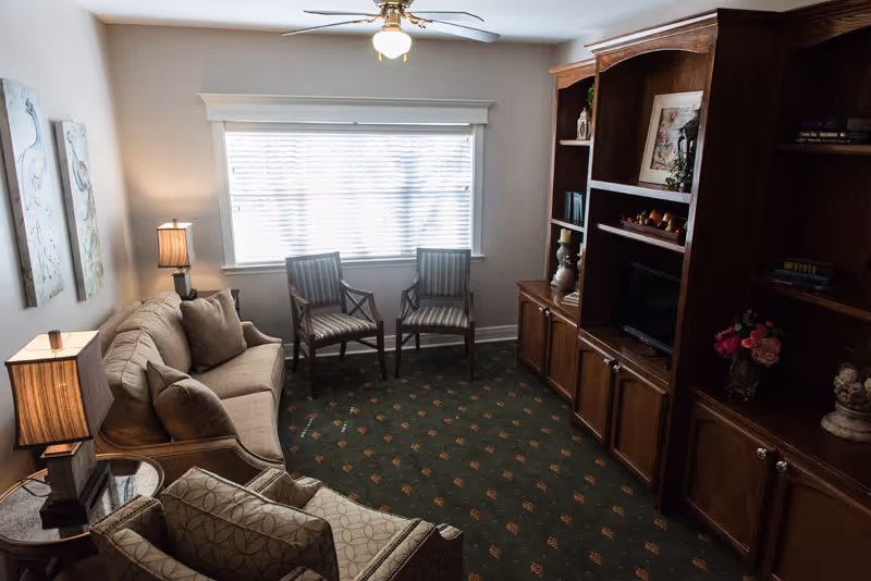 A cozy living room with a beige sofa, two wooden armchairs with striped cushions, a patterned armchair, a wooden entertainment center with shelves and a TV, two table lamps with beige shades, and a large window with blinds letting in natural light.
