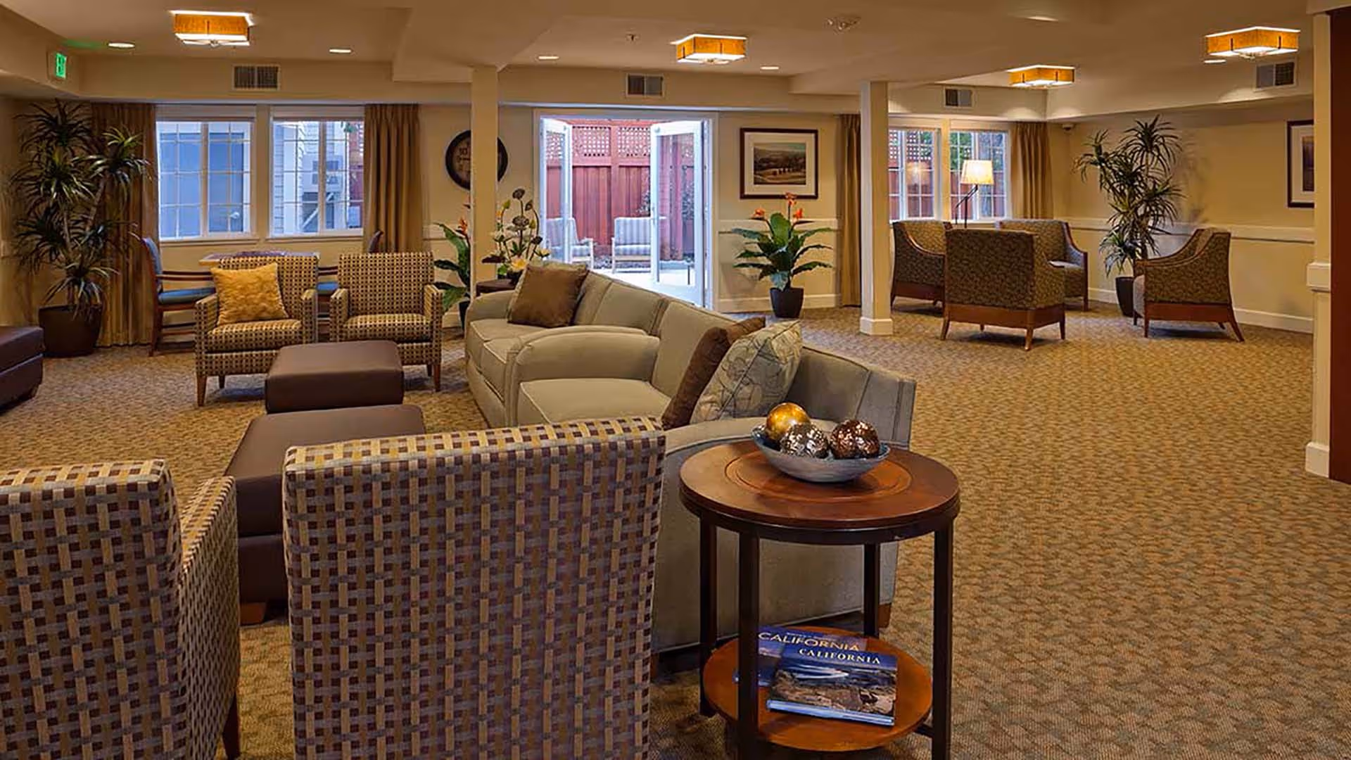 A spacious, well-lit living room area with multiple seating arrangements including sofas, armchairs, and ottomans. The room features carpeted flooring, potted plants, framed artwork on the walls, and large windows with curtains. Double glass doors open to an outdoor patio area with additional seating.