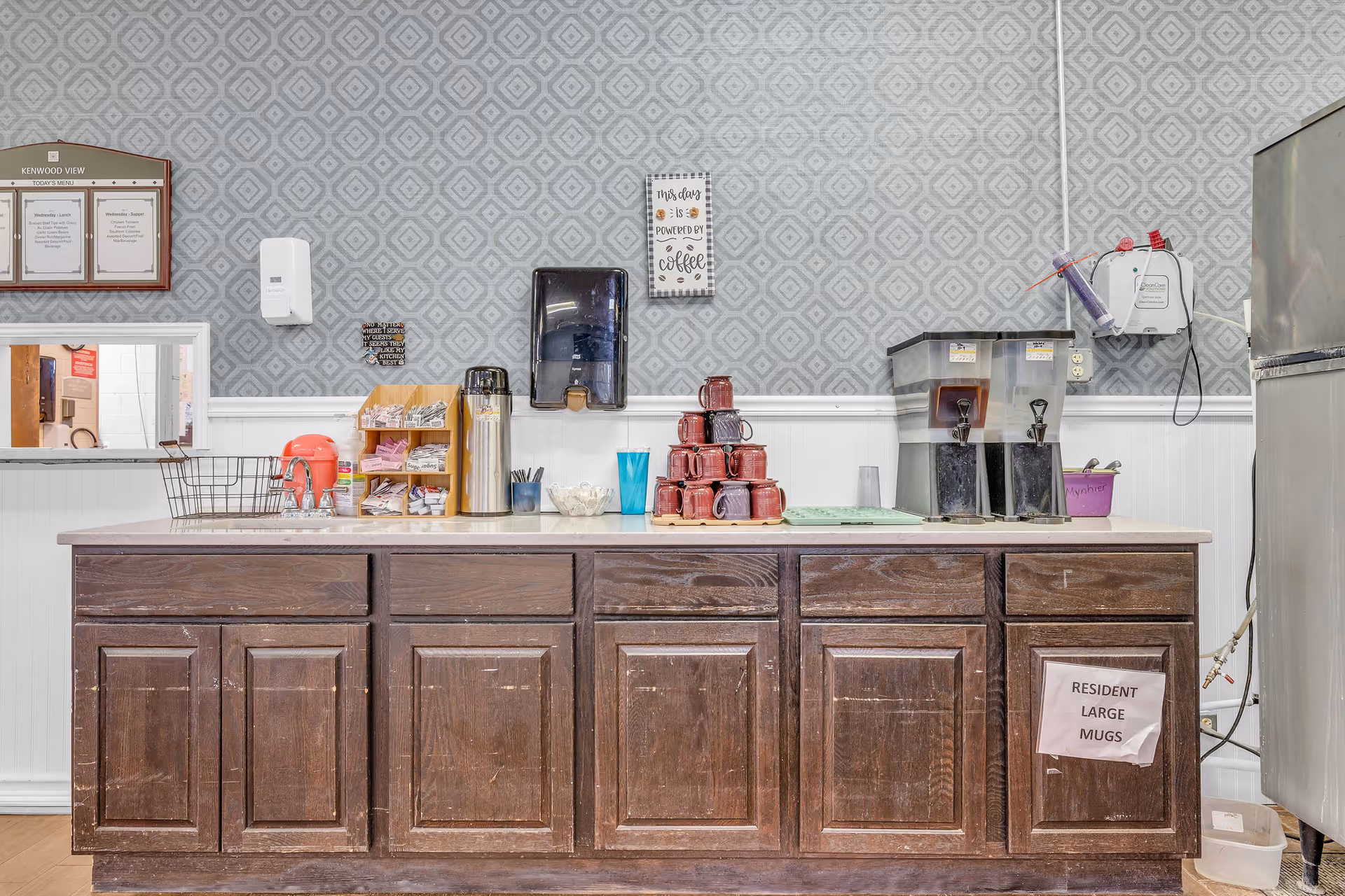 A beverage station in Kenwood View Health and Rehabilitation Center with a wooden cabinet below a countertop. On the countertop are stacked red mugs, a coffee dispenser, two large beverage dispensers, a metal basket, a red electric kettle, a stainless steel thermos, and various condiments and utensils. The wall behind has a patterned wallpaper with a paper towel dispenser, a sign that reads 'This day is powered by coffee,' and a menu board labeled Kenwood View. A sign on the cabinet reads 'Resident Large Mugs.'