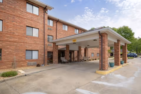 Exterior view of a brick senior living facility building with a covered entrance supported by brick columns. There are benches under the covered area and some potted plants near the entrance. The sky is partly cloudy.