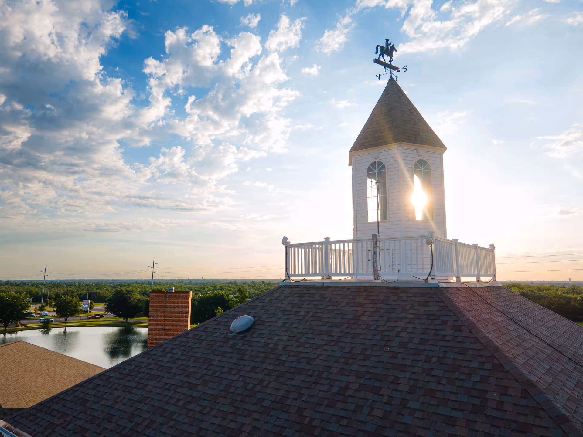 A shingled rooftop with a white cupola and weather vane, sunlight shining through its window, overlooking trees and a pond.