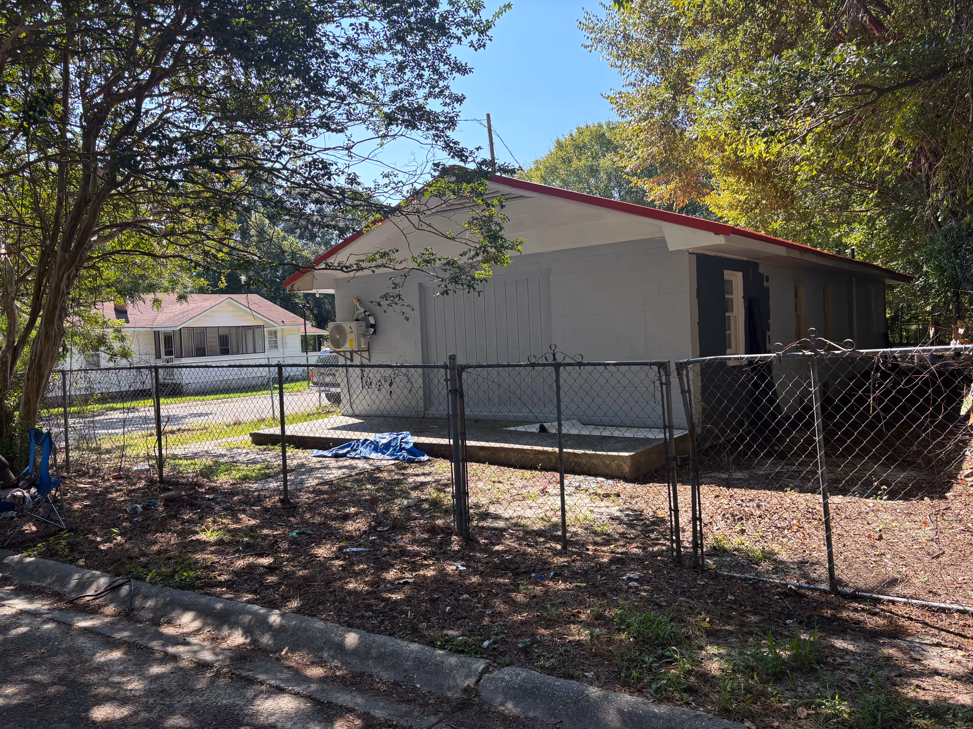 A single-story building with a red roof sits behind a chain-link fence in a shaded yard.