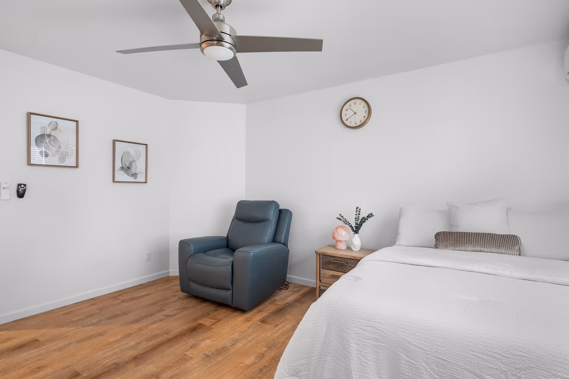 Bright minimalist bedroom featuring a bed, recliner, nightstand, clock, and ceiling fan.
