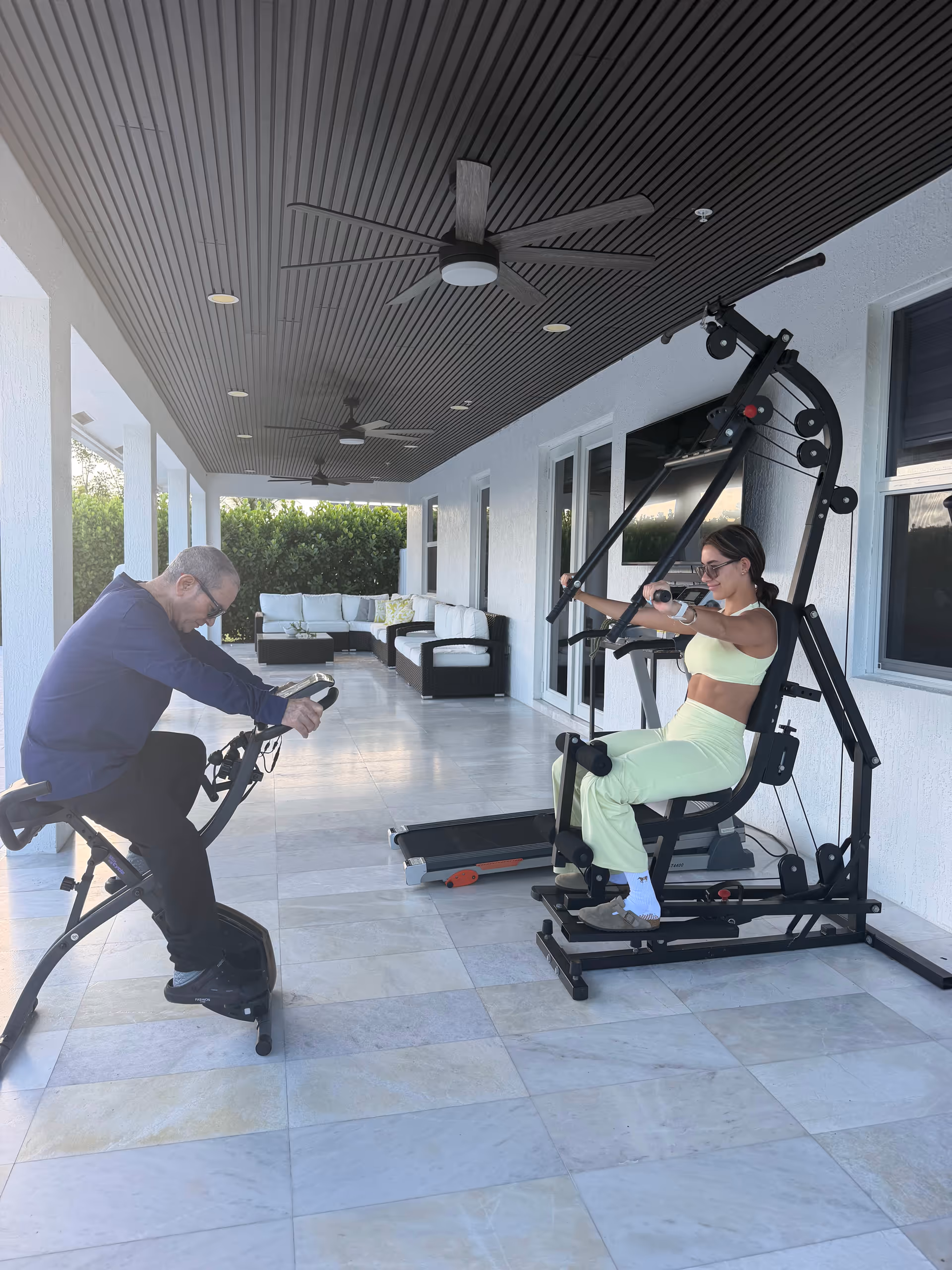 Covered outdoor patio with two adults exercising on gym equipment and a seating area.