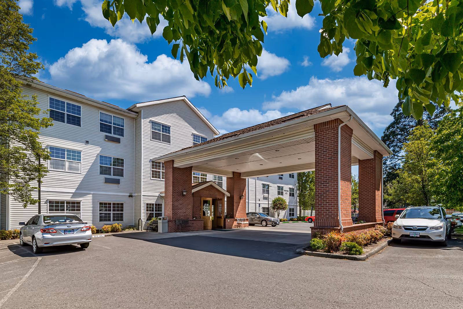 Exterior view of Courtyard At Mount Tabor senior living facility showing a three-story white building with multiple windows, a covered entrance supported by brick pillars, several parked cars, and green trees under a blue sky with scattered clouds.