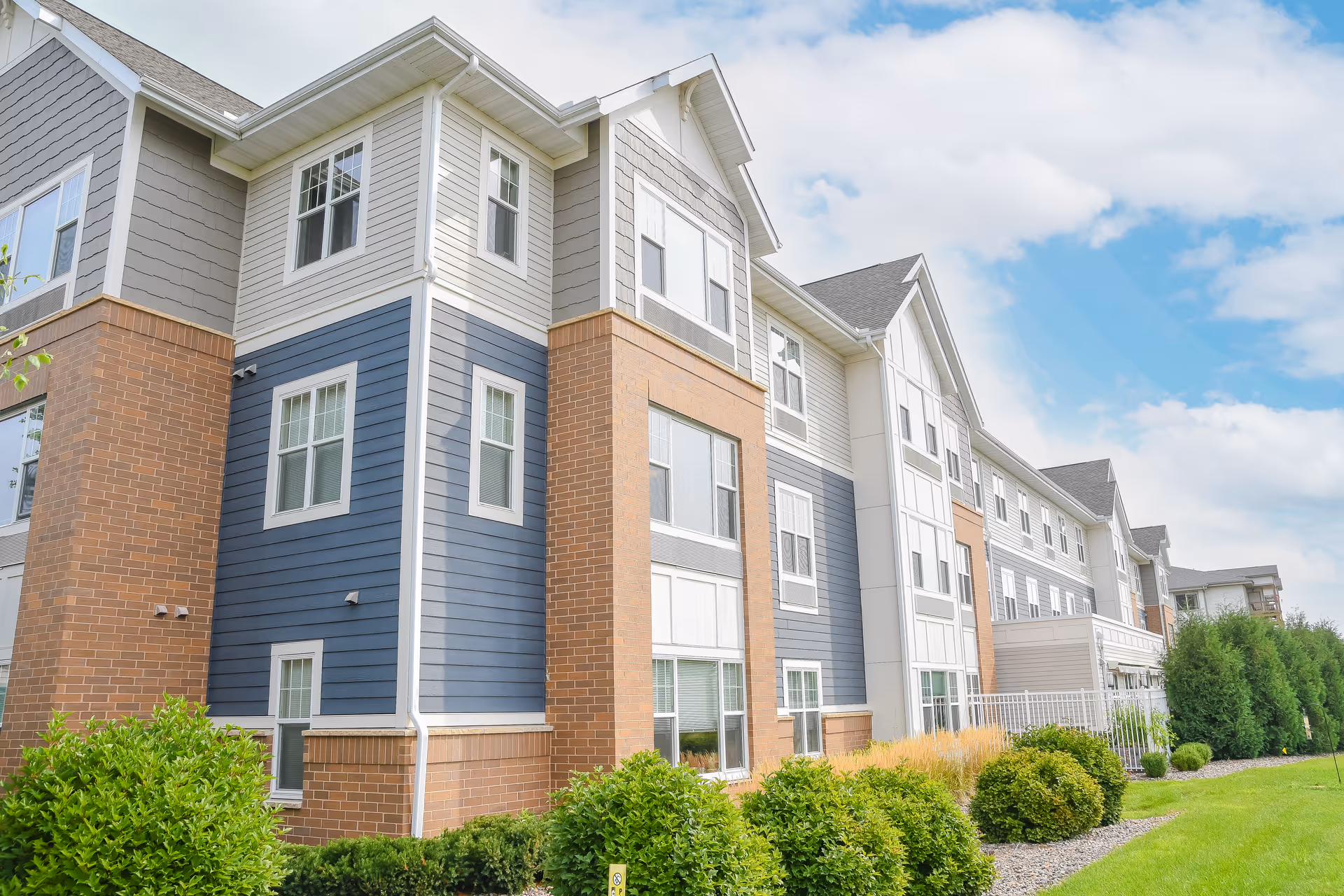 Exterior view of a multi-story senior living facility building with a combination of brick and siding in blue, gray, and white colors. The building is surrounded by green bushes and a well-maintained lawn under a partly cloudy sky.
