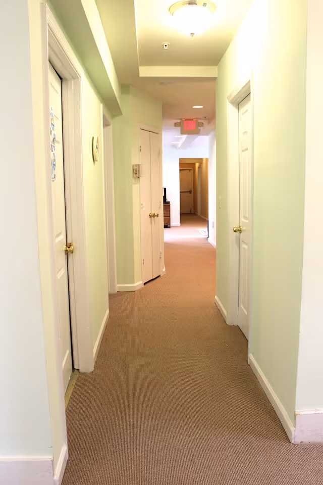 A well-lit indoor hallway with light green walls and beige carpet. There are several white doors with golden handles on both sides of the hallway. A ceiling light fixture is visible, and an exit sign is seen at the far end above a doorway.
