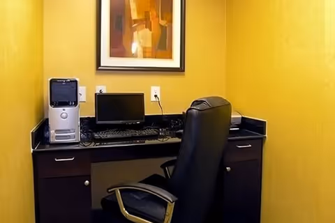 Small office workstation with a desktop computer, keyboard, black leather chair, and framed artwork on yellow walls.