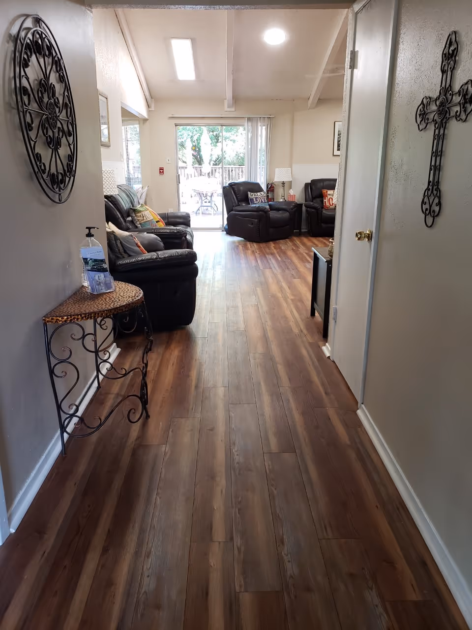 A hallway with wooden flooring leading to a living room area furnished with black leather recliners and sofas. The walls are decorated with a metal wall art piece and a decorative cross. A small table with hand sanitizer and brochures is placed against the left wall. Large sliding glass doors at the end of the room open to an outdoor patio with trees visible outside.