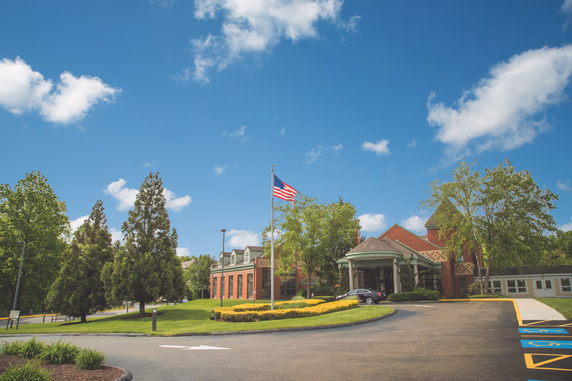 Exterior view of a senior living facility named Evergreen Woods with a circular driveway, an American flag on a flagpole, well-maintained landscaping, and a clear blue sky with some clouds.