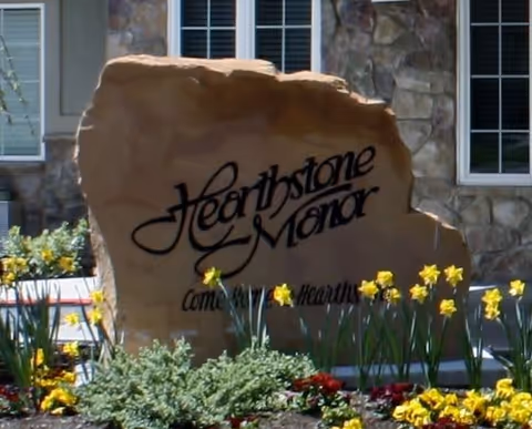 A large stone monument sign with the text 'Hearthstone Manor' and partially visible smaller text below it, surrounded by yellow daffodil flowers and greenery in front of a stone building with windows.
