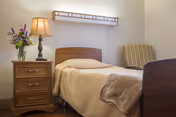 Single-bed bedroom with a wooden nightstand holding a lamp and vase of flowers, a wall light above the bed, and an upholstered chair.