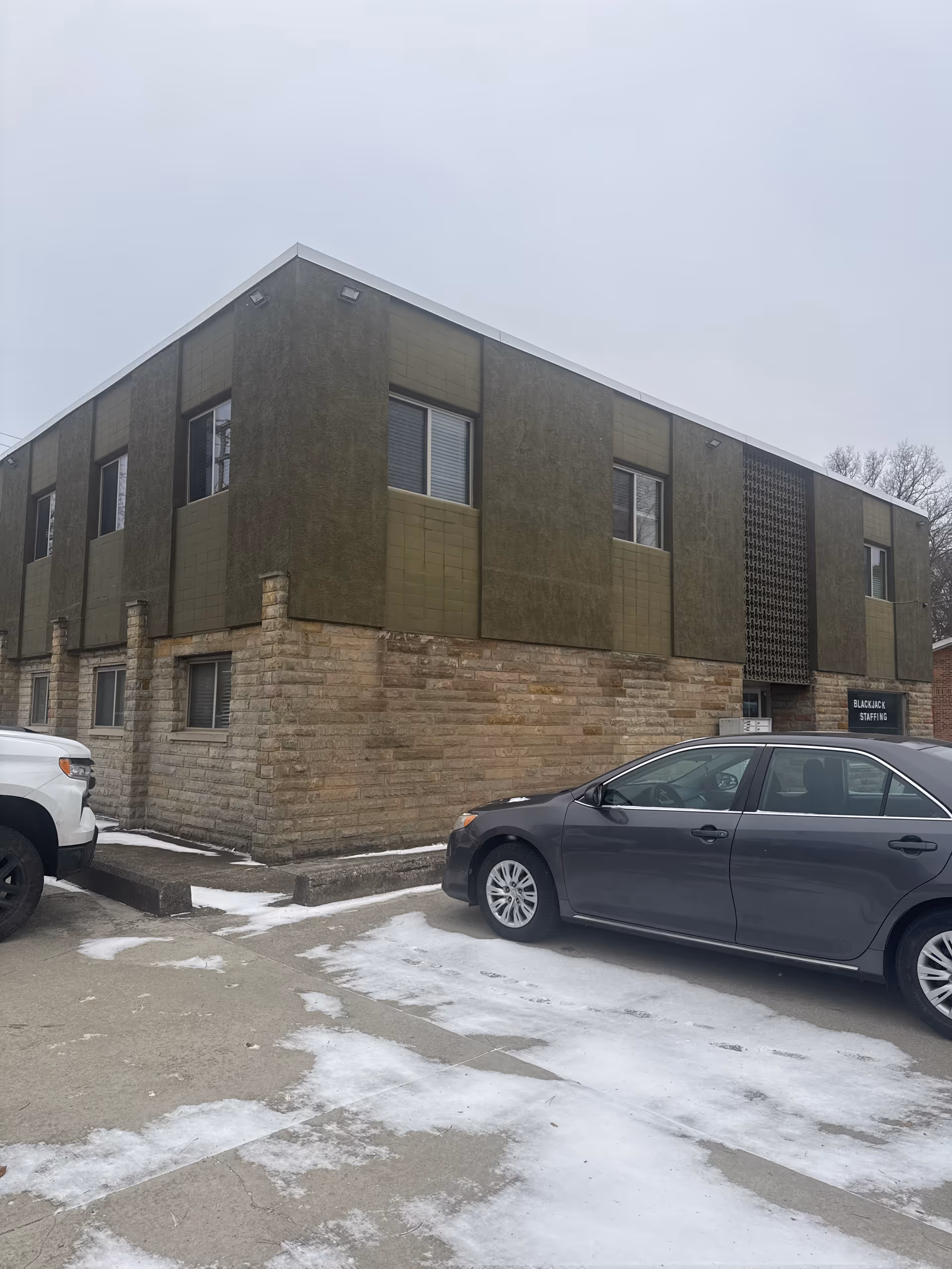 Two-story building with stone base and green panel facade, parked cars, light snow on ground