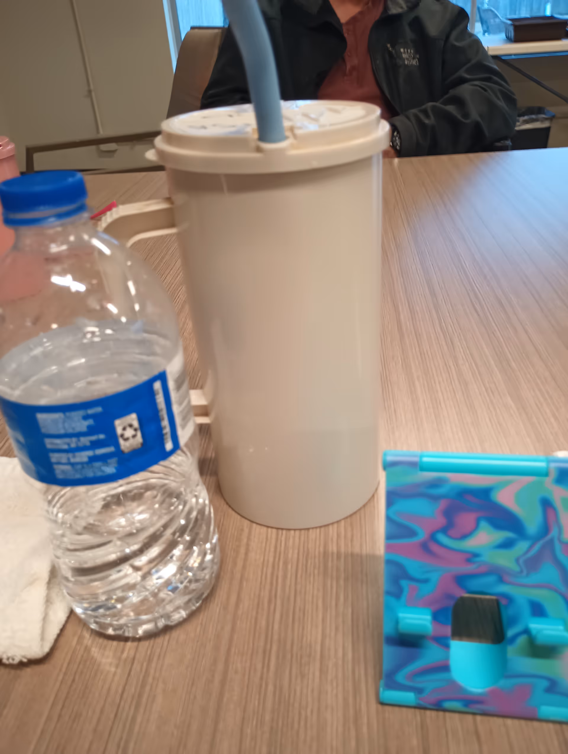 White tumbler, water bottle, and colorful case on a wooden table inside a facility.