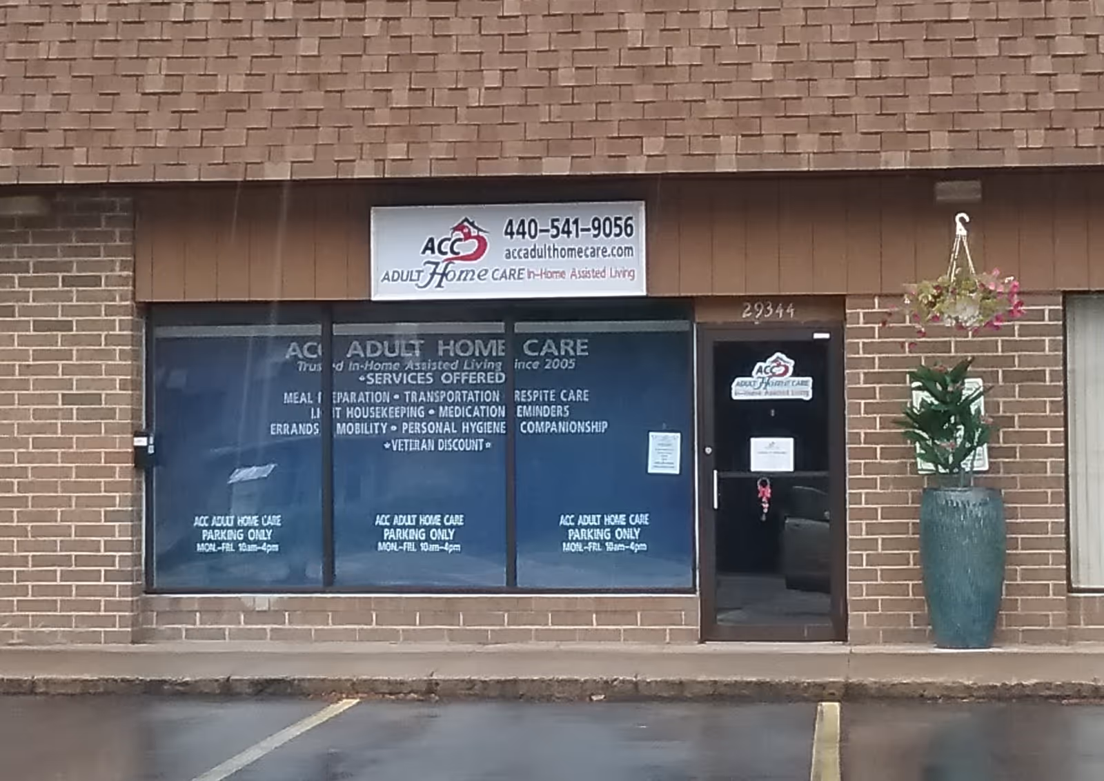 Brick storefront displaying ACC Adult Home Care signs, glass windows, and a tall potted plant.