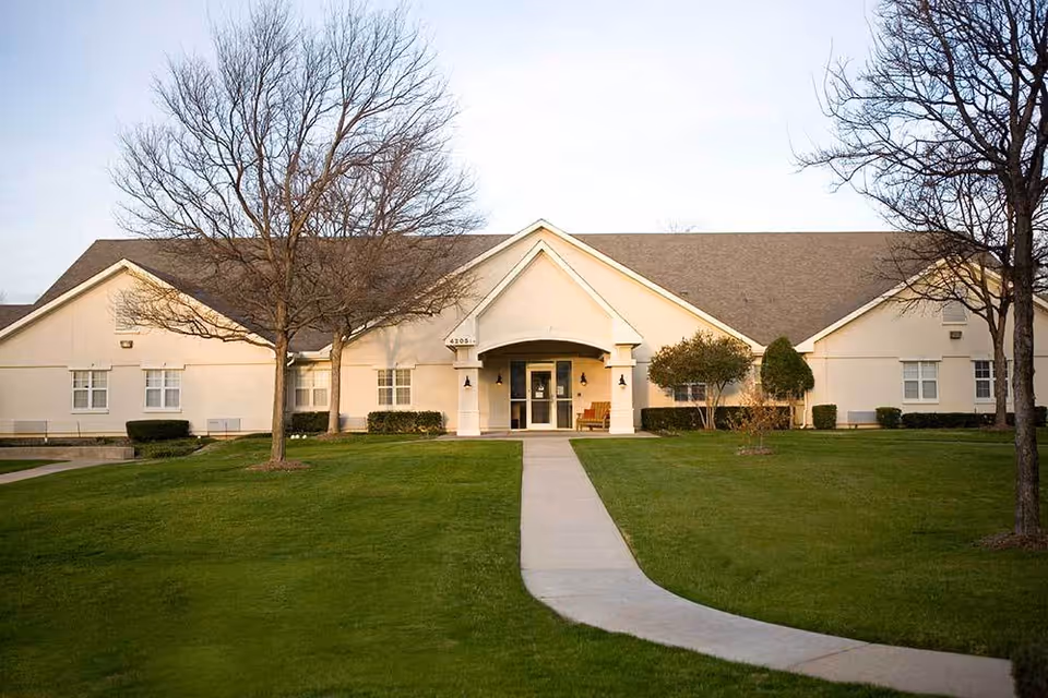 Front exterior view of a single-story building with a peaked roof, beige walls, and a central entrance with a covered porch. There is a concrete walkway leading up to the entrance, flanked by green grass and several leafless trees.