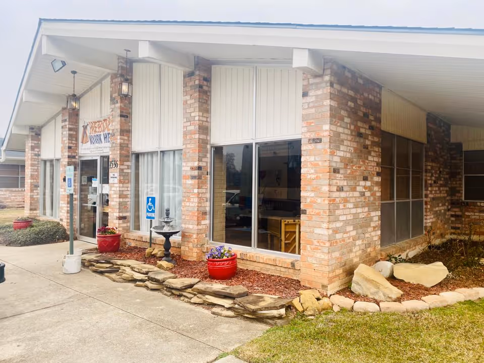 Exterior view of a brick building with large windows and a covered entrance. There are red flower pots with plants, a small stone fountain, and a handicapped parking sign near the entrance. The building has a sign above the door that reads 'Work Hub'.
