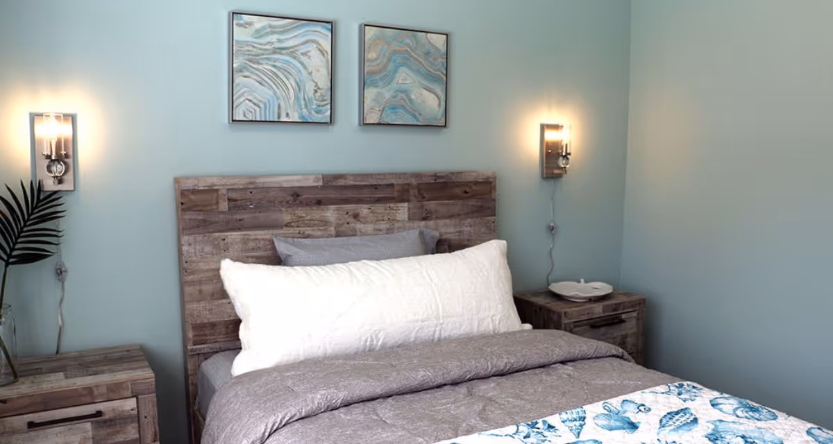 A cozy bedroom with a wooden headboard and matching nightstands on either side. The bed is made with gray bedding, a large white pillow, and a decorative blue and white quilt. Two abstract blue and gray paintings hang on the light blue wall above the bed. Wall-mounted lights are on either side of the bed, and a green plant is visible on the left nightstand.