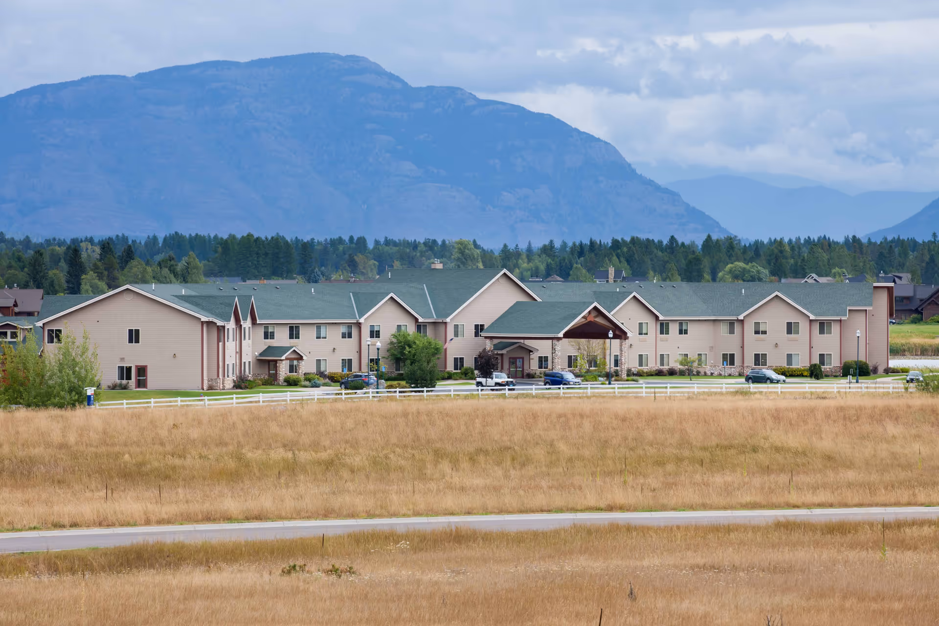 Front exterior of a long beige senior living facility with green roofs set in a grassy field and mountains in the background.