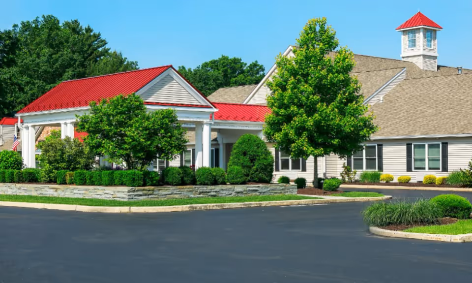 Exterior view of a senior living facility with a red-roofed entrance canopy, beige siding, multiple windows, and well-maintained landscaping including trees and shrubs under a clear blue sky.