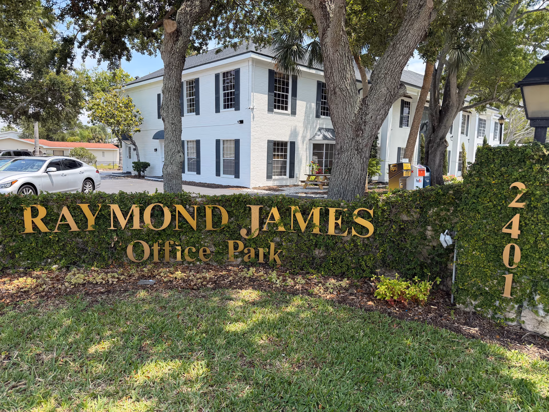 Two-story white building with blue shutters behind a hedge and a large Raymond James sign.