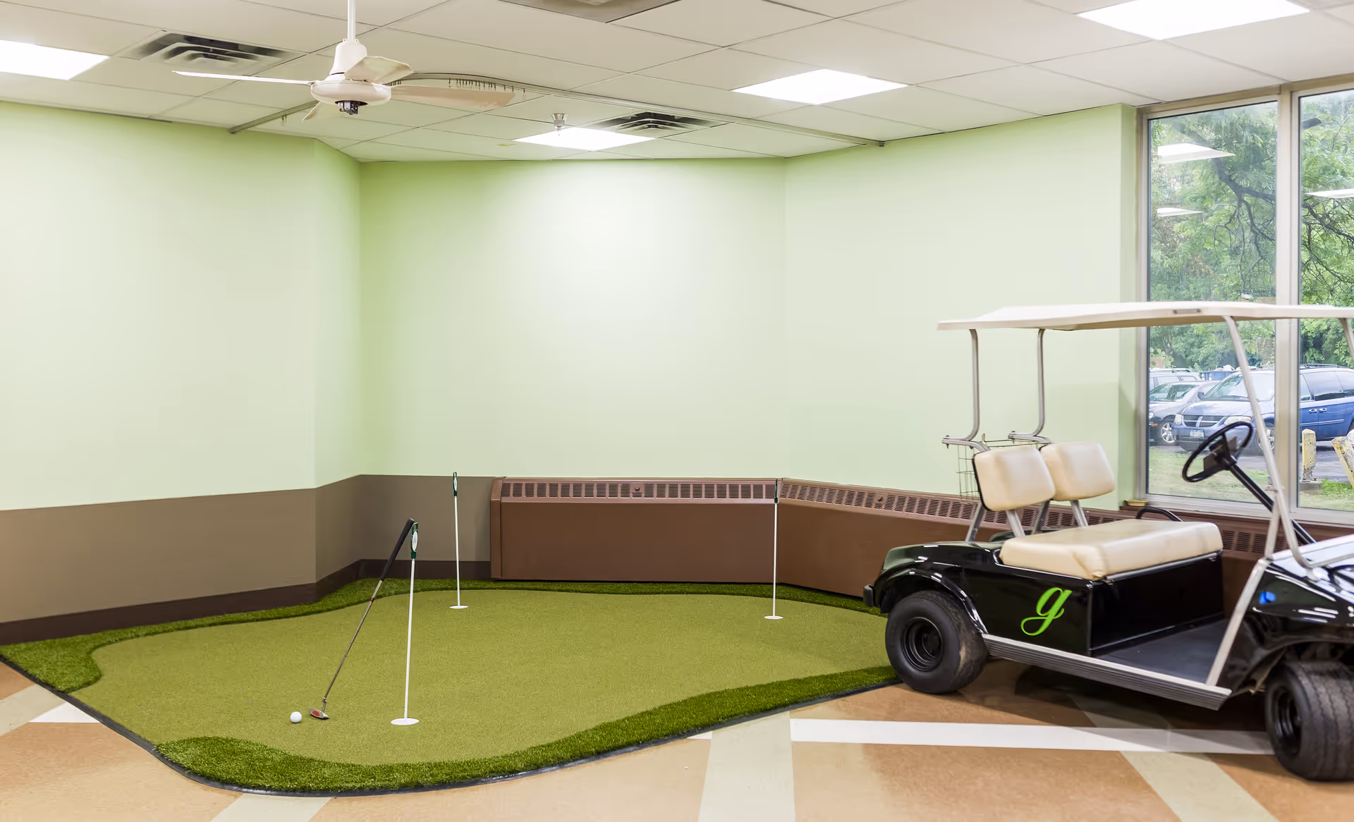 Indoor mini putting green with three holes and a parked golf cart beside a window in a pale green-walled room.