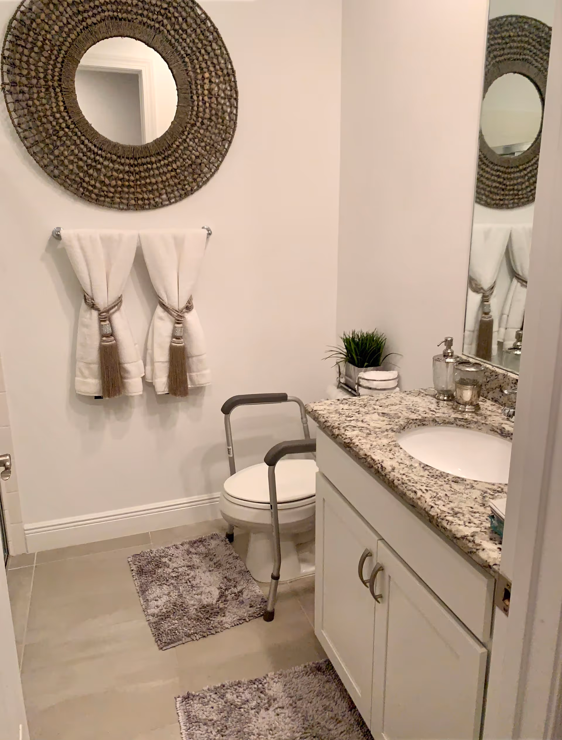 Two white tassel towels hang by a round woven mirror over a granite bathroom countertop.