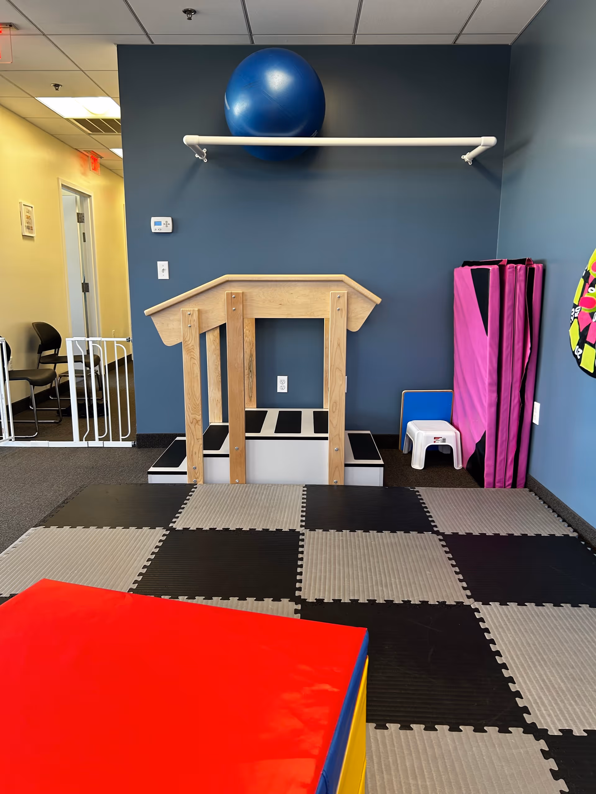 Indoor sensory gym with a wooden play frame, large blue ball, and cushioned checkerboard flooring.
