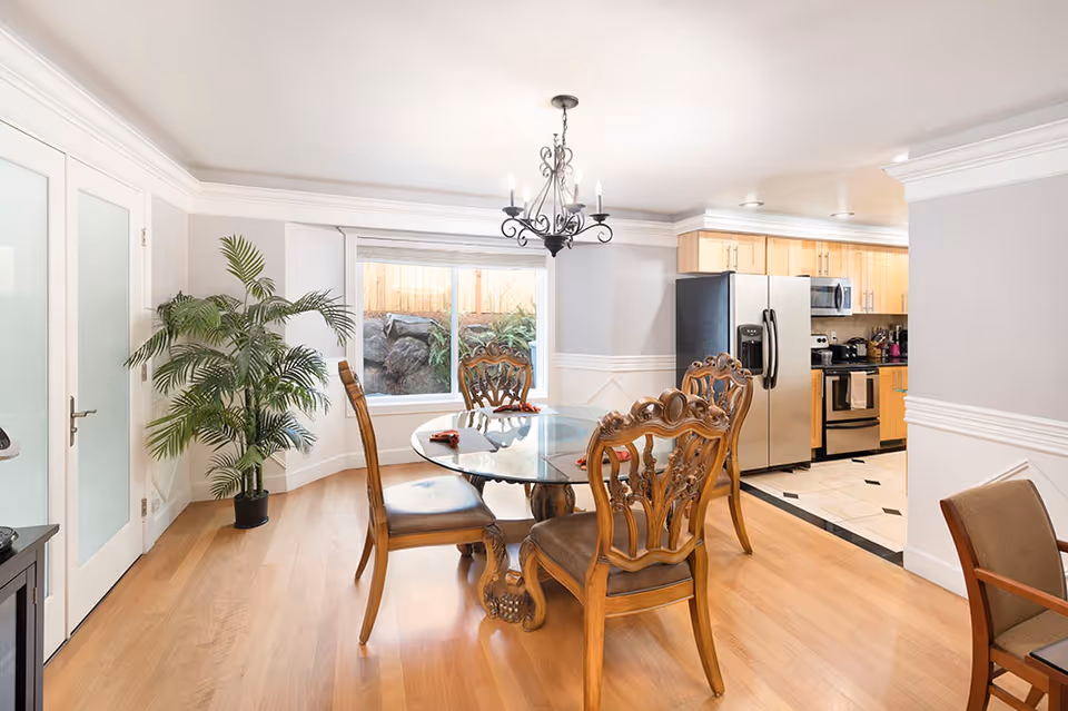 Bright dining area with glass table, ornate chairs, chandelier, plant, and adjacent kitchen.