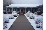 Snow-covered senior living facility front entrance with a concrete path and bushes.