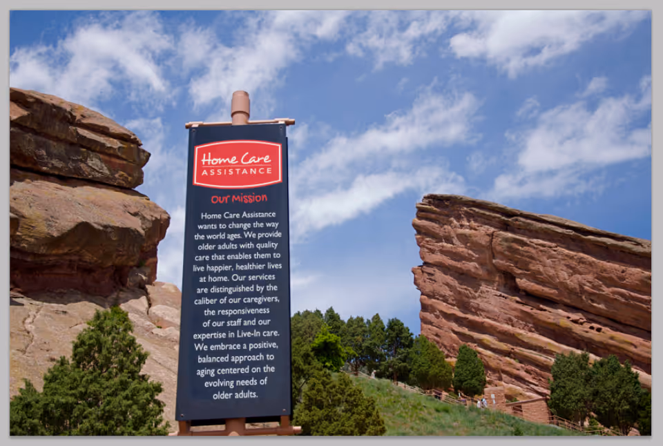 Tall signboard reading Home Care Assistance stands beside rocky hills and trees outdoors.