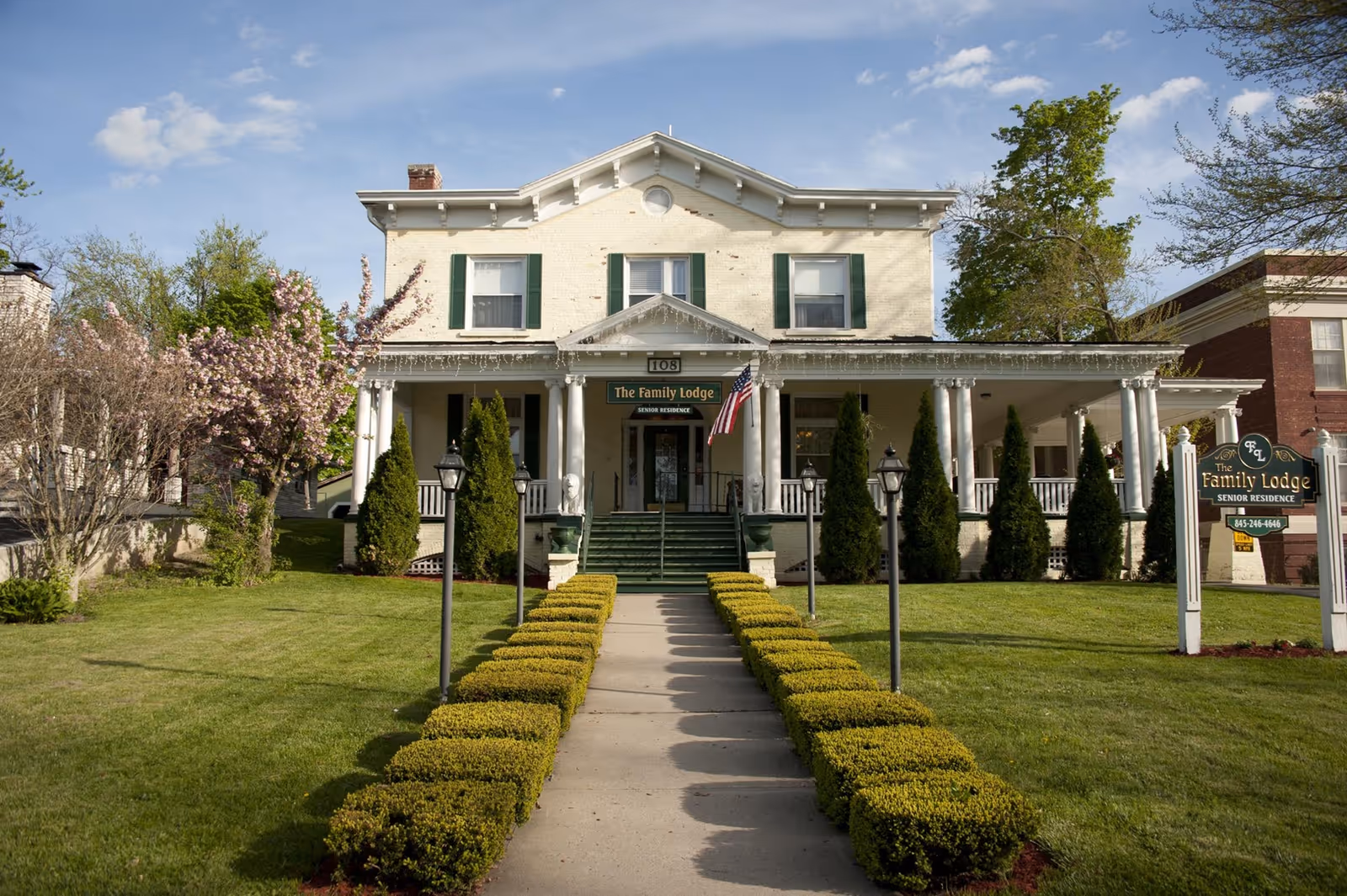 Front exterior of a two-story assisted living building with a wide porch, American flag, central walkway and trimmed hedges.