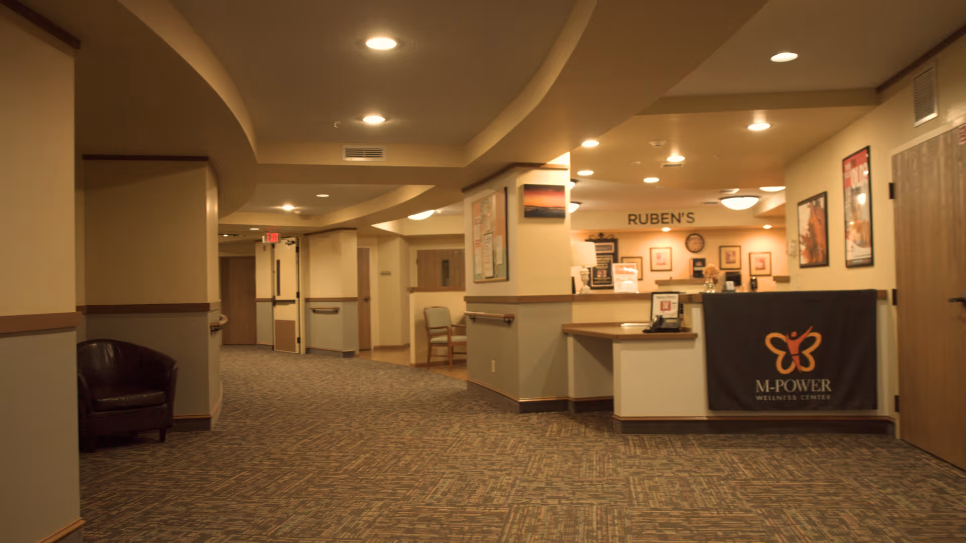 Interior hallway of a senior living facility with carpeted floor, beige walls, and ceiling lights. On the right side, there is a reception desk with a black cloth banner displaying the M-POWER Wellness Center logo. Behind the desk, a sign reads 'RUBEN'S'. There are chairs along the walls and framed pictures hanging on the walls.
