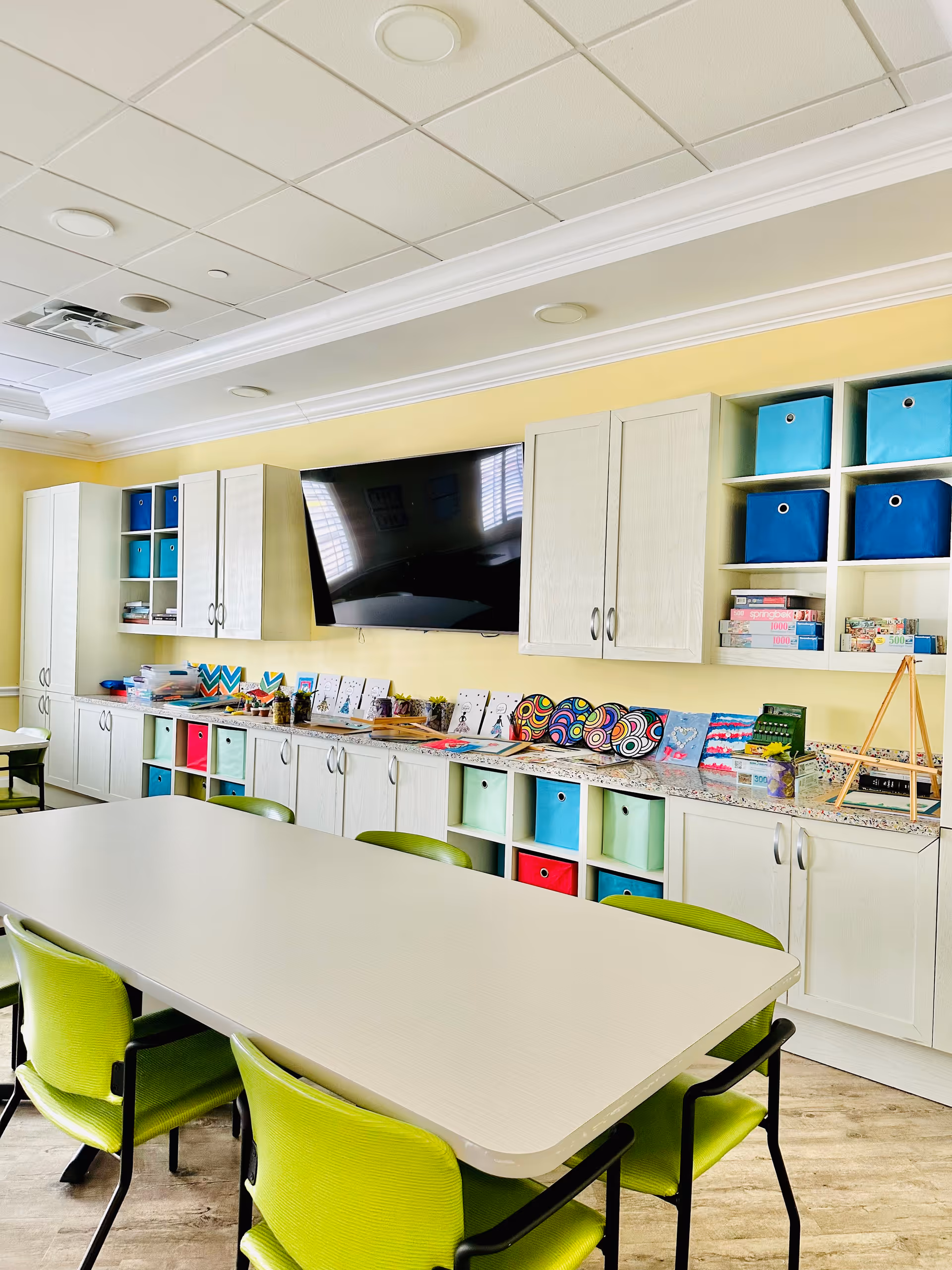Bright activity room with cabinets, colorful storage bins, and a long teaching table and green chairs.