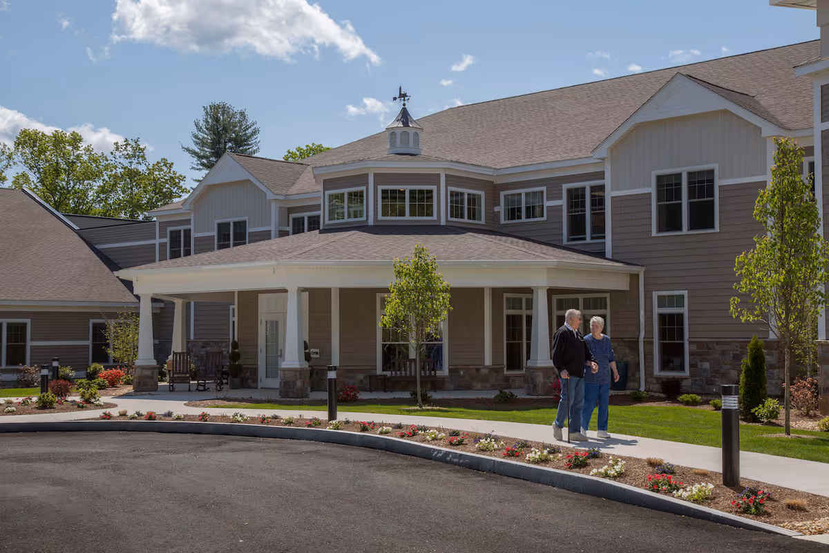 Front entrance of a senior living building with a covered portico and two elderly people walking on the sidewalk.