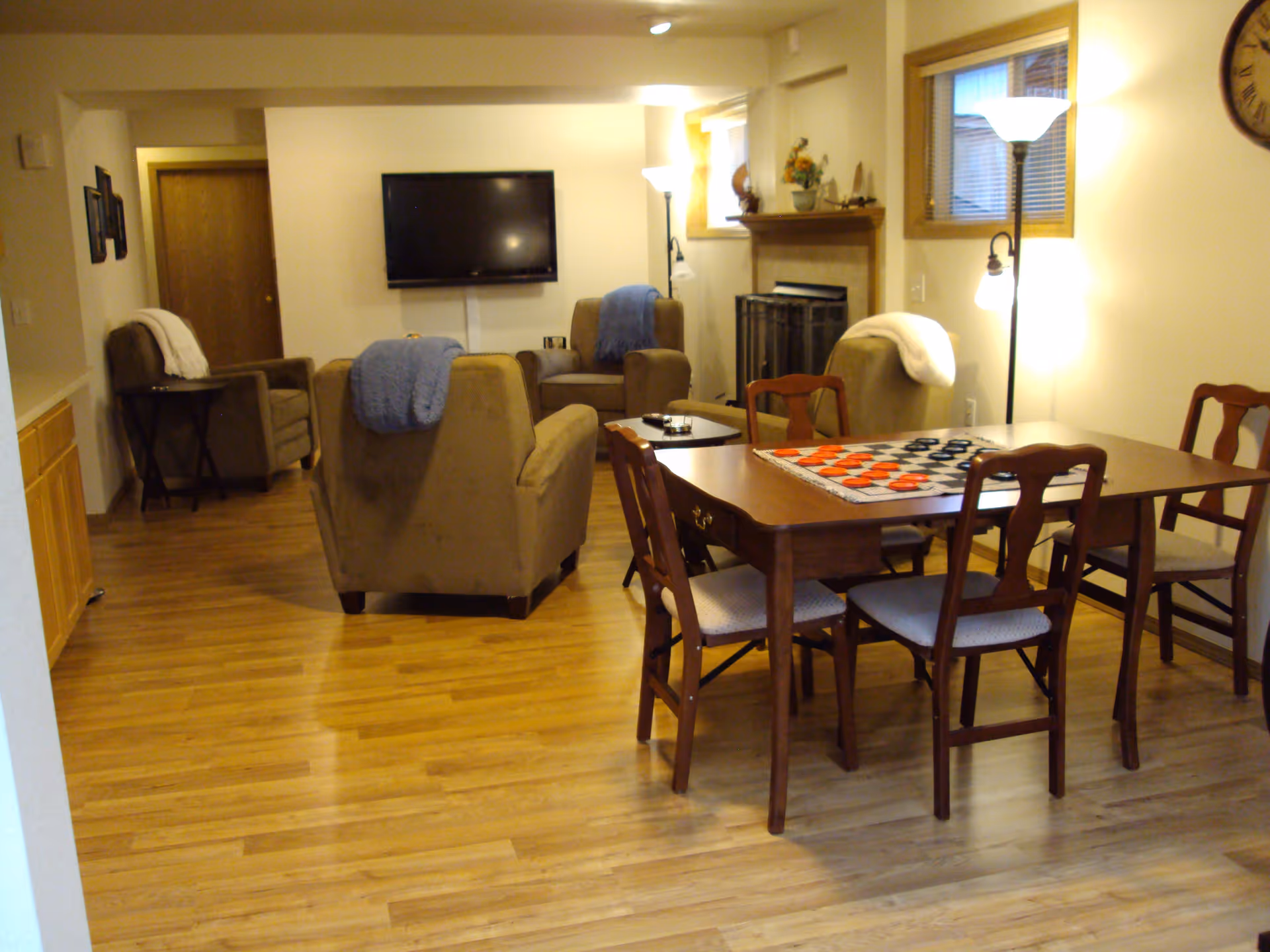 A cozy living room and dining area with wooden flooring, featuring a wooden dining table with four chairs and a checkers board on top. The living room has several cushioned armchairs with blankets draped over them, a wall-mounted flat-screen TV, a fireplace with decorative items on the mantel, and floor lamps providing warm lighting.